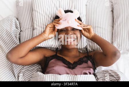 Faul fröhlich Millennial afroamerikanische Frau zieht aus Schlafmaske und schaut in die Kamera, liegt auf dem Bett Stockfoto