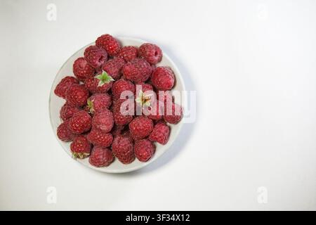 Eine Top-Down-Nahaufnahme eines runden weißen Keramikschüssels, der mit frischen reifen roten Himbeeren gefüllt ist. Die Schüssel steht auf der linken Seite einer klaren Stockfoto