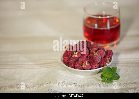 Eine horizontale Nahaufnahme einer kleinen weißen Schale gefüllt mit reifen roten Himbeeren und einem Minz Zweig auf einem gefransten Leinentischtuch. Ein Glas Rotwein Stockfoto
