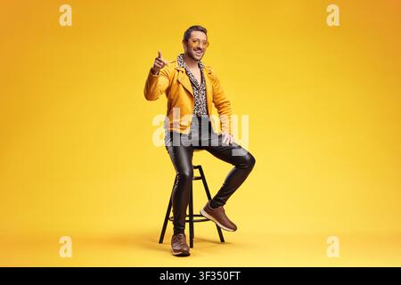 Mann in einer gelben Jacke, der auf einem Hocker sitzt und isoliert auf gelbem Hintergrund zeigt Stockfoto