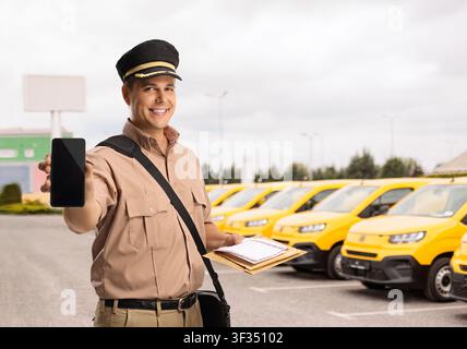 Postbote zeigt einen Smartphone-Bildschirm vor einer Flotte gelber Lieferwagen Stockfoto