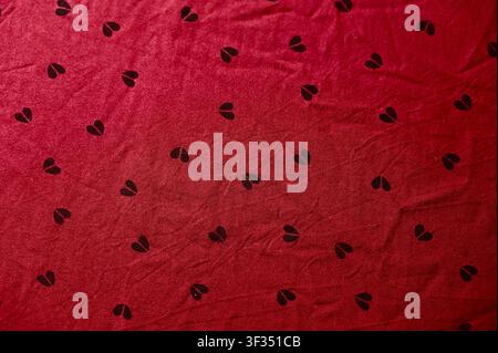Roter Stoff mit Herz Nahaufnahme von zerknittertem rotem Stoff mit kleinen schwarzen Herzdrucken. Weiche Falten und dynamische Schatten erzeugen eine gemütliche und romantische Textur. Idealer Hintergrund für Valentinstag, Liebeskonzepte, Modedesign oder Textil-Vorlagen. Hintergrund mit weichen Wellen. Hochwertiges Foto Stockfoto