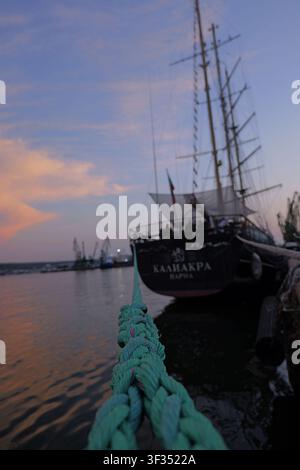 Ankerleine im Vordergrund, die auf das Segelboot Kaliakra führt, das im Hafen von Varna liegt, unter einem pastellfarbenen Sonnenuntergangshimmel mit sichtbaren Frachtkranen in t Stockfoto