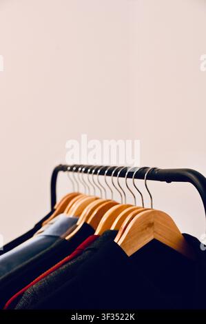 Nahaufnahme von hölzernen Kleiderbügeln mit lässiger Kleidung, die ordentlich auf einem modernen schwarzen Metall-Kleiderständer hängt, vor einem schlichten weißen Wandhintergrund. Ideal für Mode, Capsule Wardrobe, Einzelhandel, Schrankorganisation und minimalistische Lebensstilkonzepte. Stockfoto