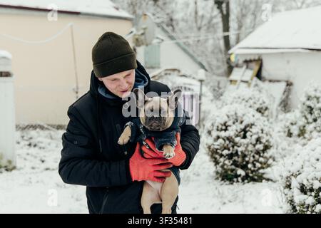 Ein Mann hält einen kleinen Hund, während er in einem verschneiten Hof steht. Der Hof ist mit weißem Schnee bedeckt, wobei einige Büsche sichtbar sind. Der Mann trägt Handschuhe und eine Ha Stockfoto