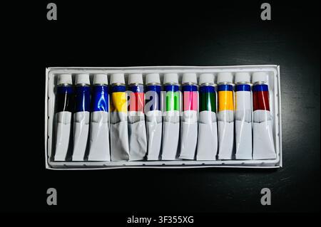 Set von farbenfrohen Farbtuben in einer Schachtel, Blick von oben auf schwarzem Hintergrund. Hochwertiger Blick von oben, flaches Layout einer offenen Schachtel mit einem farbenfrohen Set von Acryl-, Öl- oder Wasserfarbtuben, die in einer Reihe auf einem dunklen, texturierten Hintergrund angeordnet sind. Perfektes Konzept für Kunstbedarf, Kreativität, Malhobby, Schulerziehung und künstlerische Designprojekte. Stockfoto