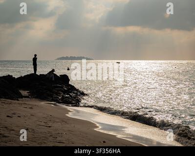 Menschen, die auf schroffen Küstenfelsen am glitzernden Meer stehen und sitzen, während sie den Sonnenuntergang vom Strand in Karimunjawa, Jepara, In beobachten Stockfoto