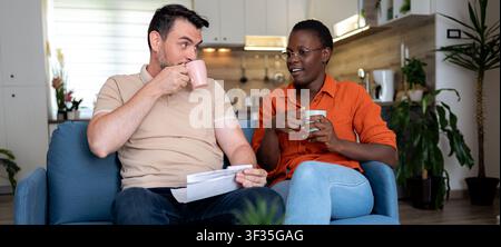 Ein Paar sitzt auf einer blauen Couch in ihrem Zuhause, nippt an Getränken und unterhält sich. Sie befinden sich in einem gemütlichen Küchenbereich mit Pflanzen um sie herum. Stockfoto