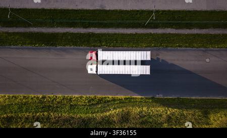 Riga, Lettland - 16. Juli 2026: Luftaufnahme von zwei Sattelzügen, die entlang einer Straße fahren, die von grünem Gras und Feldern gesäumt ist. Stockfoto
