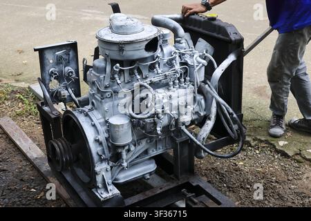 Ein alter Dieselmotor auf Metallrahmen mit freiliegenden Schläuchen, Kraftstoffpumpe und Kühler, die im Freien inspiziert werden, ein Mechaniker arbeitet in der Nähe mit lässiger Kleidung Stockfoto