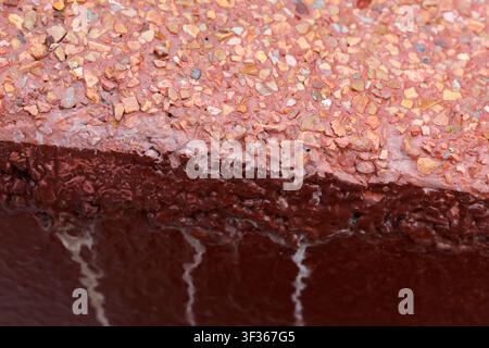 Eine rote texturierte Betonkante mit freigelegtem Zuschlag und rostfarbener Farbe, Nahaufnahme einer verwitterten Oberfläche, die körnige Steine und weiße Streifen zeigt Stockfoto