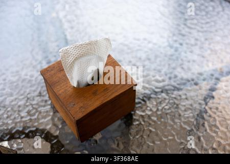 Eine Holztissuebox auf einem strukturierten Glastisch mit einer herausgezogenen weißen Papiertücherrolle, natürliches Licht und warme Holzmaserung schaffen eine gemütliche, alltägliche Szene Stockfoto