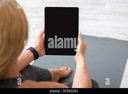 Heimtraining mit Geräten. Erwachsene Frau mit Fitness-Tracker und Tablet sitzt auf der Matte Stockfoto