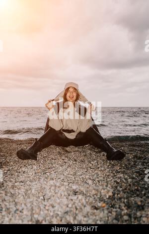 Frau am Strand mit Uschanka, verspielte Frau sitzt auf einem Kiesstrand, trägt einen Winterhut und streckt die Zunge heraus an einem bewölkten Tag Stockfoto