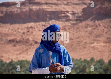 Tafilalet-Palmenhain, Marokko, 30. Mai 2026, Ein Mann in einem traditionellen blauen Berber-Tunika und Turban posiert in einer Mittelaufnahme vor der atemberaubenden n Stockfoto