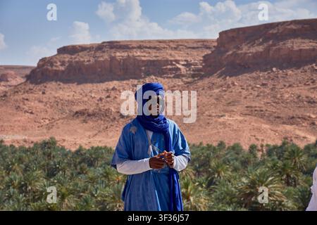 Tafilalet-Palmenhain, Marokko, 30. Mai 2026, Ein Mann in einem traditionellen blauen Berber-Tunika und Turban posiert in einer Mittelaufnahme vor der atemberaubenden n Stockfoto