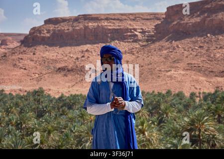 Tafilalet-Palmenhain, Marokko, 30. Mai 2026, Ein Mann in einem traditionellen blauen Berber-Tunika und Turban posiert in einer Mittelaufnahme vor der atemberaubenden n Stockfoto