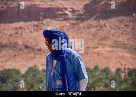 Tafilalet-Palmenhain, Marokko, 30. Mai 2026, Ein Mann in einem traditionellen blauen Berber-Tunika und Turban posiert in einer Mittelaufnahme vor der atemberaubenden n Stockfoto