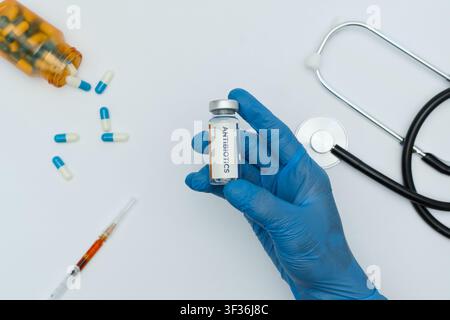 Handschuh tragende Hand hält eine Impfampulle mit Stethoskop und Pillen auf weißem Hintergrund. Stockfoto