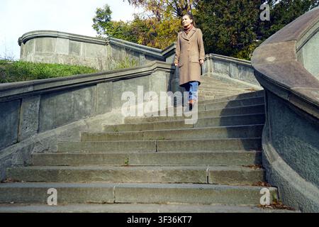 Frau in beigem Wollmantel steigt breite Steintreppe im herbstlichen Stadtpark hinab. Zeigt alltägliche urbane Mobilität, ruhige saisonale Bewegung, Geh-Routine Stockfoto