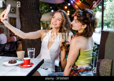 Zwei junge Frauen lächeln und posieren für ein Selfie an einem Cafétisch. Sie genießen Getränke und Snacks in einer lebhaften Außeneinrichtung. Stockfoto