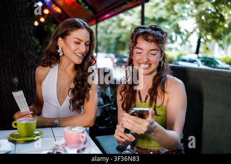 Zwei Frauen sitzen an einem Tisch in einem Café. Sie lächeln und schauen auf ein Telefon, während sie ihre Getränke genießen. Stockfoto