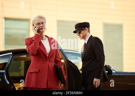 Senior weiße Geschäftsfrau telefoniert mit dem Smartphone, während sie aus dem Auto steigt, wobei der Chauffeur wartet, und vermittelt Führungsposition und den Einsatz von Premium-Service für Corporate Branding Stockfoto