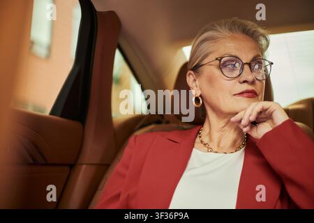 Mittlereuropäische Geschäftsfrau mittleren Alters sitzt im Auto, schaut durch das Fenster und reflektiert während der Fahrt. Ideal für Führungskräfte, Unternehmensreisen, Erfolgsmarketing Stockfoto