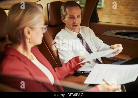 Erfahrene kaukasische Geschäftsfrau und Geschäftsmann, die Dokumente durchsehen, während sie im Auto sitzen, und über Vertragsbedingungen und Geschäftszusammenarbeit diskutieren. Für geschäftliche Reisen, Abschluss von Verträgen, Führungsteam-Zusammenarbeit Stockfoto
