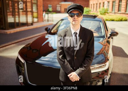Porträt eines kaukasischen Chauffeurs mittleren Alters vor einer Luxuslimousine, mit Sonnenbrille, der Premium-Transportdienst und geschäftliche Reisen vermittelt. Für Gastgewerbe, Geschäftsmobilität-Marketing Stockfoto