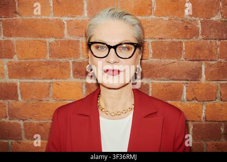 Porträt einer älteren kaukasischen Geschäftsfrau mit Brille, die in die Kamera schaut vor einer Ziegelwand, die Selbstvertrauen und Führung vermittelt. Ideal für Führungskräfte-Branding und Karrieremarketing Stockfoto