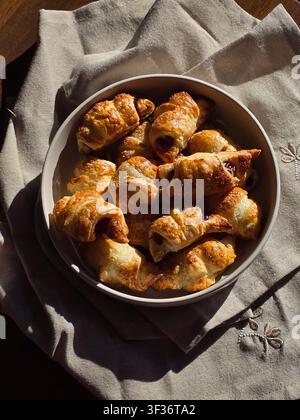 Frisch gebackene Mini-Croissants mit Marmeladenfüllung auf einem Keramikteller serviert. Helles natürliches Sonnenlicht erzeugt dramatische Schatten auf einer Leinenserviette. Herbst Stockfoto