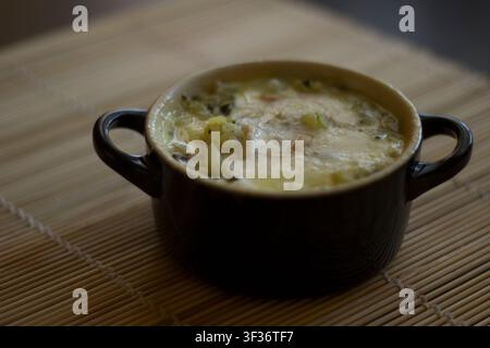 Dunkle Keramikcocotte mit zwei Henkeln, gefüllt mit gebackenem Zucchini, Kartoffeln und Pinienkernen unter geschmolzenem Toma-Käse, auf einer Bambusmatte, bei schwachem Licht Stockfoto