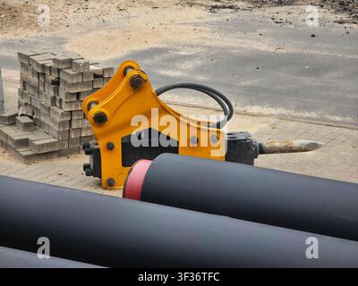 Gelber hydraulischer Hammeraufsatz auf dem Boden. Große schwarze Industrierohre, Stapel von grauen Pflastersteinen sichtbar. Szene zeigt aktive Baustelle Stockfoto