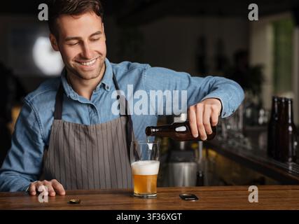 Der Barkeeper gießt Bier aus der Flasche in Glas und steht hinter der Theke im Inneren Stockfoto