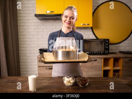 Lachende Frau hält einen frisch gebackenen runden Kuchen auf einem Tablett in einer modernen Küche mit gelben Schränken Stockfoto