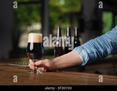 Entspannen Sie sich im Pub. Barkeeper Hand legte auf hölzerne Theke ein Glas dunkles Bier mit Schaum Stockfoto