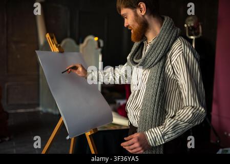 Bärtiger Künstler, der an einer Staffelei in warmem Studiolicht mit strukturierter Wand und Kunstmaterialien skizziert Stockfoto