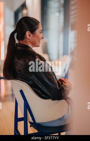 Junge Frau in Lederjacke, die in einem modernen Bürostuhl sitzt und im Profilblick lächelt Stockfoto