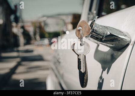 Makro-Nahaufnahme eines Sets von Vintage-Autoschlüsseln, die im Chromtürschloss eines klassischen Autos hängen, mit unscharfem Straßenhintergrund Stockfoto