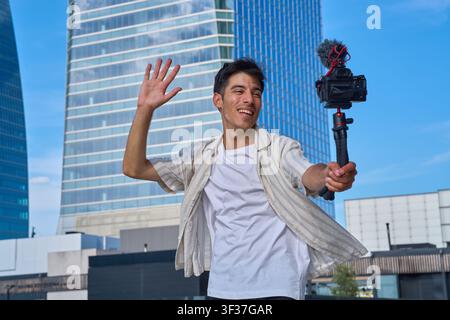 Junger Mann hält eine Kamera mit einem Stativ, winkt, während er im Freien in einer modernen Stadt Video aufnimmt Stockfoto