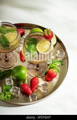 Zwei elegante Glascocktails mit Minzblättern, Limettenstücken und Eiswürfeln, serviert neben frischen Erdbeeren und ganzen Limetten auf einem metallischen Servierteller Stockfoto
