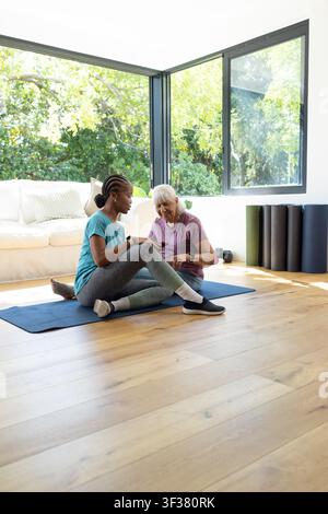 Zwei Frauen, Seniorin und Trainerin in Sportkleidung, sitzen auf blauer Matte zu Hause und überprüfen ein Wearable Stockfoto