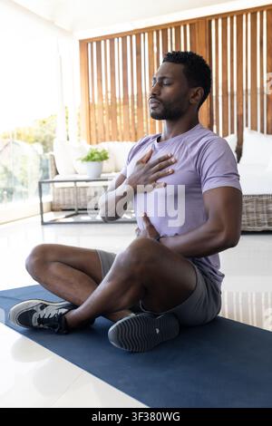 Afroamerikaner sitzt auf blauer Yogamatte in einem Wintergarten und trägt ein lila Hemd und graue Shorts Stockfoto