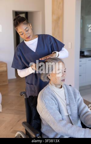 Afroamerikanische Pflegerin in OP-Kleidung, die das Haar einer älteren asiatischen Frau im Rollstuhl zu Hause glättet Stockfoto