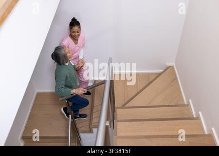 Seniorin und Pflegekraft in rosa OP-Kittel, die eine Treppe hinabsteigen und sich an einem Metallgeländer festhalten Stockfoto