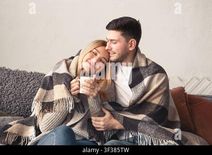 Glückliches junges kaukasisches Paar kuschelt unter der Decke zu Hause und genießt ein heißes Getränk Stockfoto