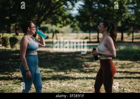 Zwei Frauen machen eine Pause vom Outdoor-Fitness und plaudern, während sie im Park Wasser trinken Stockfoto