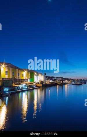 Blick auf die Uferpromenade in der Dämmerung zeigt farbenfrohe Gebäude mit leuchtenden Lichtern. Das Wasser spiegelt diese Lichter wider und erzeugt eine lebendige Szene. Boote liegen am Dock Stockfoto