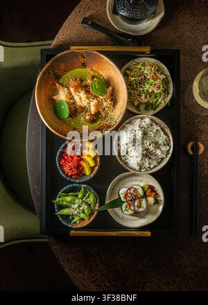 Top-Down-Ansicht eines japanischen Mittagessens auf einem schwarzen Tablett mit einer Schüssel grüner Curry, Maki-Sushi-Rollen, Reisschüssel, Edamame und frischem Salat. Stockfoto